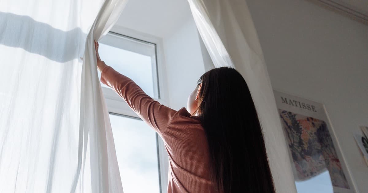 A woman seen from behind opens a light curtain in the morning, daylight falling into the room