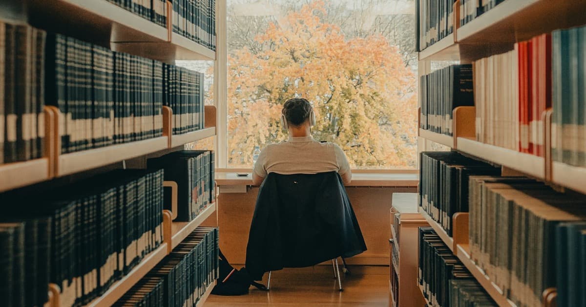 Person wearing headphones sitting by a window at the end of a library aisle